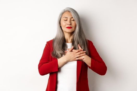 Senior asian woman with red lips and blazer, close eyes and holding hands on heart thankful, feeling nostalgic, dreaming of something, standing over white background.