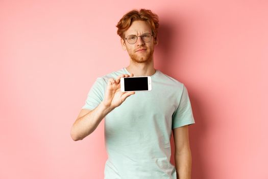 Skeptical redhead guy in glasses showing smartphone screen horizontally, smirk and frowning disappointed, standing over pink background.