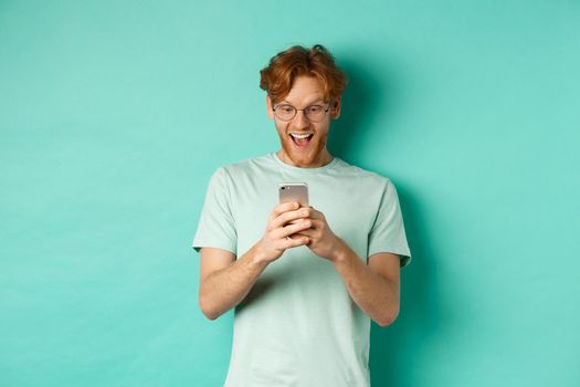 Image of young redhead man in glasses reading phone screen with surprised face, receive amazing promo offer, standing over turquoise background.