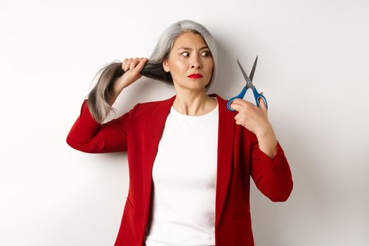 Indecisive asian woman holding scissors and looking doubtful, thinking to cut hair, changing haircut, standing over white background.