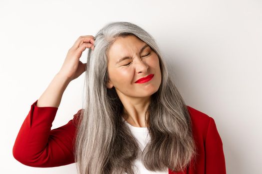 Close up of senior asian woman scratching head and looking bothered by itching, standing over white background.