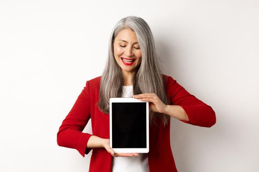 Business. Successful asian businesswoman in red blazer showing blank digital tablet screen, looking down with pleased smile, white background.