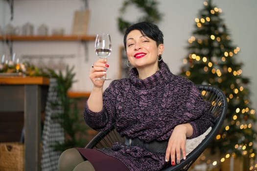 Portrait of a short hair brunette woman with a glass of champagne sits on a chair celebrating christmas.