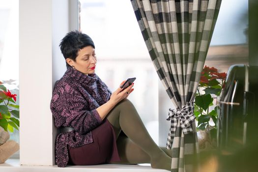 Short hair brunette woman reads news on mobile phone while sitting on windowsill.