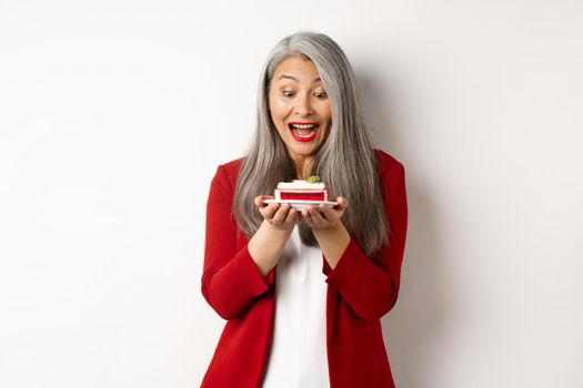 Cheerful asian businesswoman enjoying office party, looking at delicious piece of cake and smiling, standing over white background.