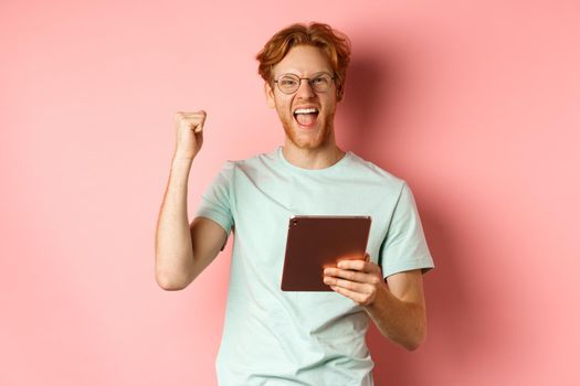 Image of happy redhead man triumphing, winning online with digital tablet and rejoicing, standing over pink background.