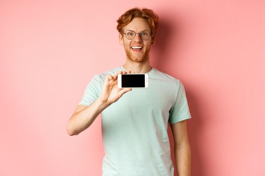 Happy young man with red hair showing smartphone screen, holding phone horizontally and smiling amazed, standing over pink background.