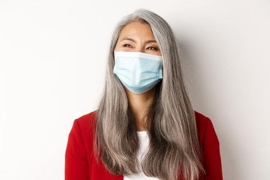 Covid, pandemic and business concept. Close up of happy asian businesswoman with grey hair, wearing medical mask and smiling, looking left with cheerful face, white background.