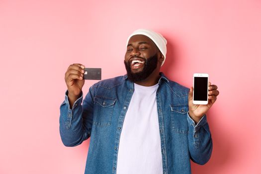 Online shopping. Happy african-american man in beanie laughing, showing credit card and mobile phone screen, standing over pink background.