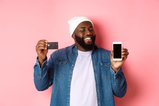 Online shopping. Satisfied Black man nod in approval, smiling and looking at phone, showing credit card and smartphone screen, standing over pink background.