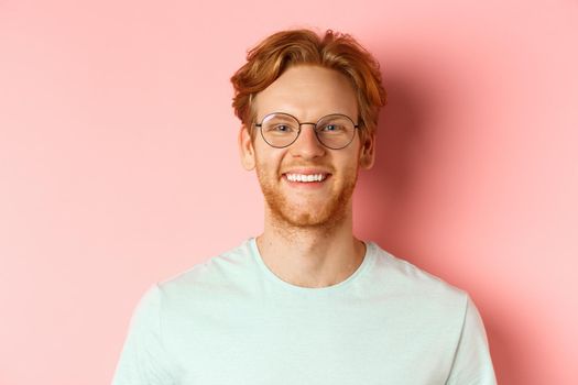 Close up of happy redhead man face, smiling with white teeth at camera, wearing glasses for better sight and t-shirt, standing over pink background.
