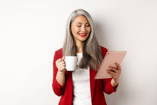 Business people concept. Elegant asian woman drinking coffee and reading on digital tablet, standing over white background.