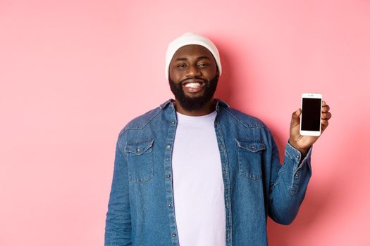 Handsome hipster guy in beanie and denim shirt smiling, showing mobile phone screen with happy face, introduce application, standing over pink background.