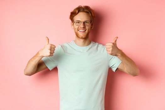 Cheerful european man with red hair and beard, wearing glasses, showing thumbs-up and smiling in approval, praise something good, standing over pink background.