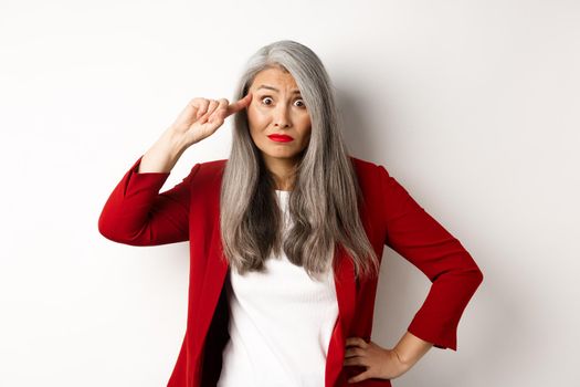 Annoyed asian business woman scolding someone stupid, showing crazy gesture, pointing finger at head and staring at camera, white background.