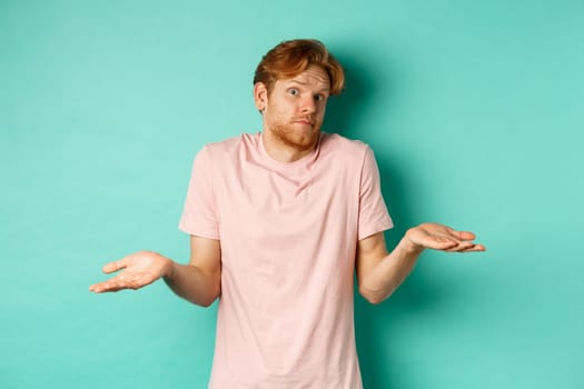 Young redhead guy in t-shirt know nothing, shrugging shoudlers with hands spread sideways, standing confused over turquoise background.