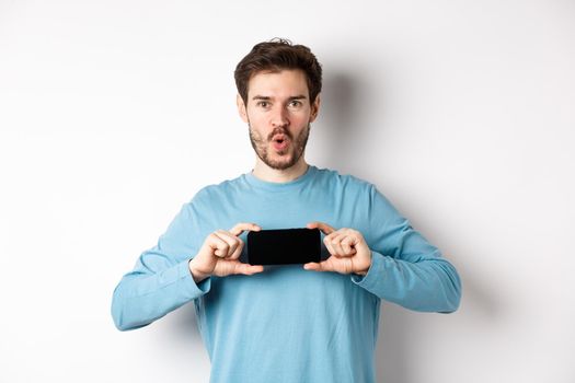 E-commerce and shopping concept. Image of impressed man saying wow and showing blank mobile phone screen in horizontal, standing over white background.