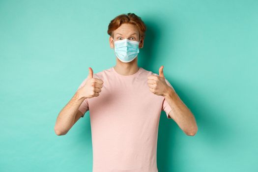 Covid-19, pandemic and lifestyle concept. Cheerful redhead guy in medical mask showing thumbs up in approval, like and praise product, standing over turquoise background.