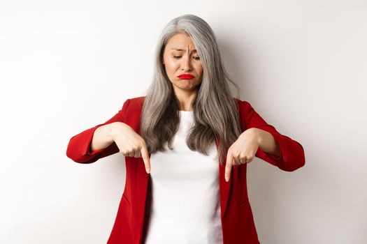 Sad and gloomy asian senior woman in red blazer sulking, pointing fingers down and looking upset, standing over white background.