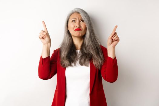 Disappointed asian senior woman looking, pointing fingers up and grimacing, dislike something, standing skeptical over white background.