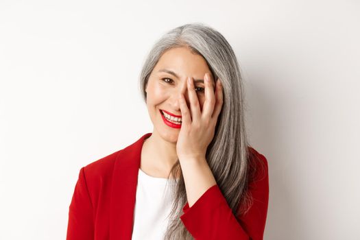 Beauty and makeup concept. Close up of beautiful asian mature woman, laughing and touching face, smiling happy, standing over white background.