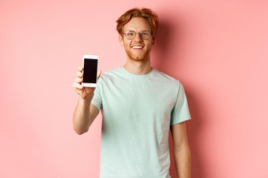 Handsome redhead man in glasses showing blank smartphone screen and smiling, demonstrate online promo or application, standing over pink background.