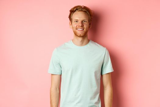 Happy redhead man in t-shirt looking at camera and smiling, standing over pink background.