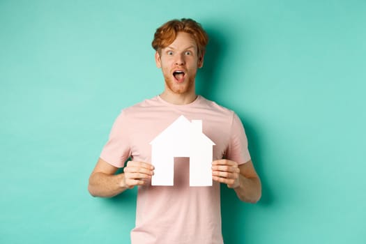 Real estate concept. Excited redhead guy with beard, showing paper house cutout and gasping in awe, staring fascinated at camera, standing over mint background.