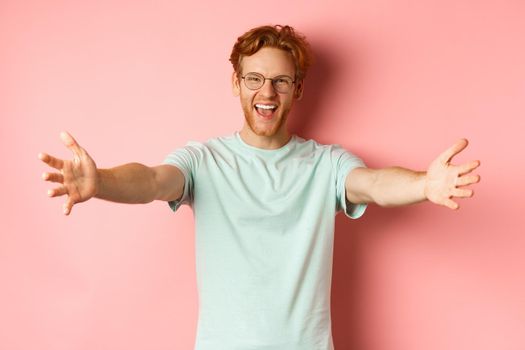 Young friendly man with red hair and beard reaching hands for hug, stretch out arm in warm welcome, smiling happily, standing over pink background.
