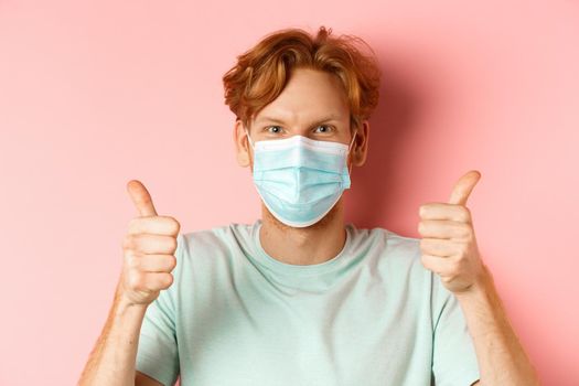 Covid-19 and pandemic concept. Handsome guy with messy ginger hair, wearing medical mask on face and showing thumbs up, standing over pink background.
