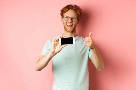 Happy european man with ginger hair and glasses, showing mobile blank screen horizontally and thumbs-up, recommend online promo, pink background.