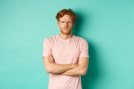 Young man with ginger hair and beard looking offended, feeling insulted and staring at camera, sulking at you, cross arms on chest defensive, standing over turquoise background.
