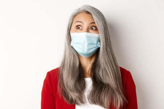 Covid, pandemic and business concept. Close up of elegant asian senior lady staring at upper left corner, wearing medical mask from coronavirus, white background.