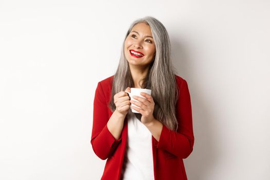 Business people concept. Smiling asian businesswoman having coffee break, holding warm mug and looking upper left corner dreamy, white background.