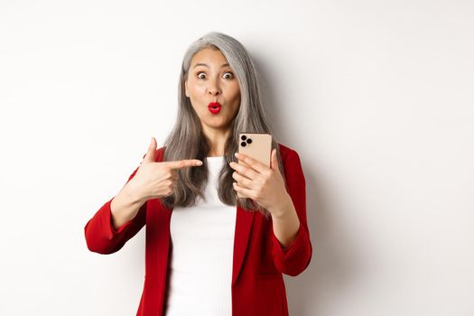 Amazed korean lady in red blazer pointing finger at mobile phone, looking curious at camera, white background.