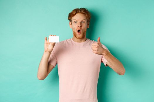 Adult guy with red hair and beard showing plastic credit card and thumb up, looking impressed, recommend bank, standing over mint background.