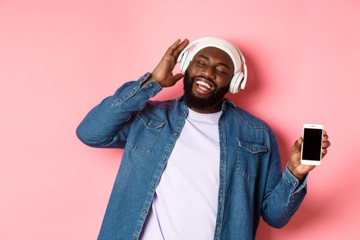 Happy african-american hipster guy listening music in headphones, showing phone screen app and singing along, standing over pink background.