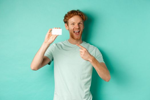 Shopping concept. Handsome redhead man in t-shirt showing plastic credit card and smiling, standing over turquoise background.