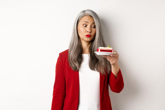Elegant senior woman in red blazer wants take bite of sweet cake, looking with tempted face at dessert, standing over white background.