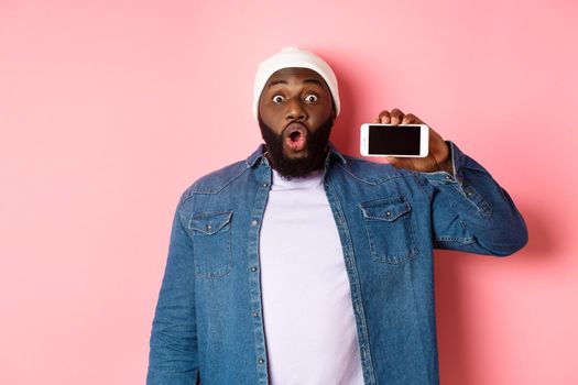 Online shopping and technology concept. Amazed Black man showing phone screen horizontally and staring at camera excited, standing over pink background.