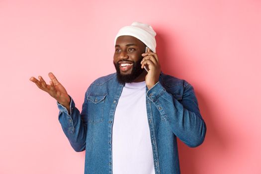 Handsome modern african-american man talking on mobile phone, smiling and discussing something, standing over pink background.