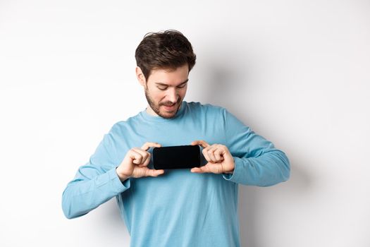 E-commerce and shopping concept. Caucasian man showing blank mobile screen and looking at it, standing curious against white background.