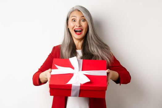 Surprised and thankful asian grandmother receiving gift in red box, looking amazed and grateful, standing over white background.