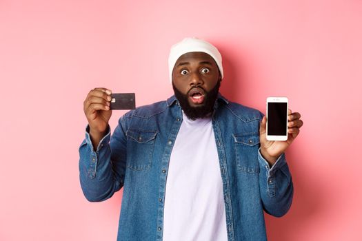 Online shopping. Shocked and concerned Black man staring at camera, showing mobile phone screen and credit card, standing over pink background.