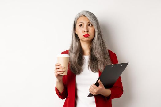 Tired asian female office worker holding clipboard and paper cup, drinking coffee and exhaling with exhausted face, standing over white background.