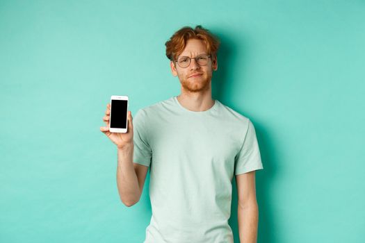 Skeptical male model with red hair and glasses, showing mobile screen and frowning disappointed, dislike application, standing over turquoise background.