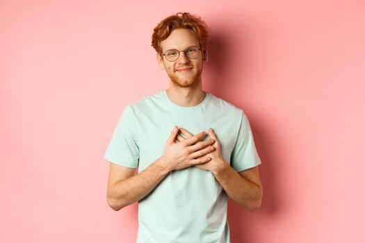 Attractive young man with ginger hair, holding hands on heart and smiling grateful, saying thank you, express gratitude, standing over pink background.