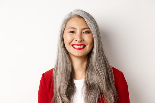 Close-up of elegant mature woman in red blazer and makeup, smiling happy at camera, standing over white background.