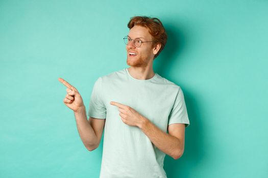 Handsome young man with red hair and beard, wearing glasses and t-shirt, pointing and looking left with amused face, checking out advertisement on copy space, mint background.