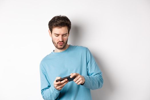 Young man playing video games on smartphone, tilt body and tap mobile screen, standing joyful over white background.